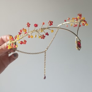 Könnte beinhalten: Ein goldener Draht-Haarreif mit einem zarten Blumenmuster aus roten und gelben Perlen. Der Haarreif hat eine herabhängende Kette mit einer roten Perle am Ende.