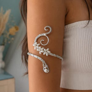 May include: Silver arm cuff with a floral design. The cuff wraps around the upper arm and features small, white flowers and a pearl accent. The design includes a spiral and a curved arm band.