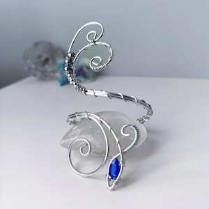 Könnte beinhalten: Ein silberfarbenes Drahtarmband mit Spiraldesign und einem blauen Edelstein. Das Armband ist um einen klaren, abgerundeten Stein gewickelt. Das Design weist mehrere Wirbel und eine Reihe kleiner Silberperlen auf.