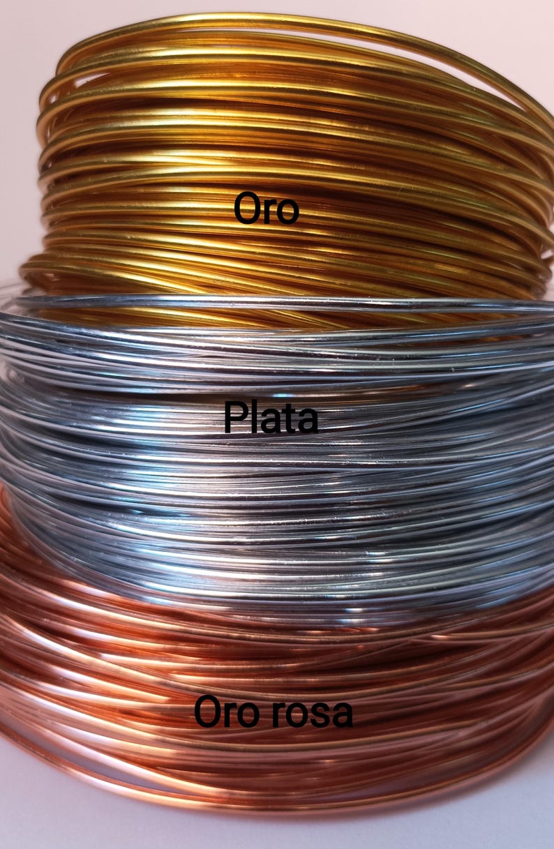K&ouml;nnte beinhalten: Drei gestapelte Spulen aus d&uuml;nnem Metalldraht in den Farben Gold, Silber und Ros&eacute;gold. Die goldene Spule ist mit dem Text Oro beschriftet, die silberne Spule mit dem Text Plata und die ros&eacute;goldene Spule mit dem Text Oro rosa.