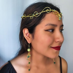 K&ouml;nnte beinhalten: Ein Kopfschmuck aus goldenem Draht mit zarten Schn&ouml;rkeln und kleinen blauen Kristallen, kombiniert mit einem langen, h&auml;ngenden Ohrring aus goldenem Draht, einer hellgr&uuml;nen marmorierten Perle und kleineren gr&uuml;nen Kristallen in gestufter Anordnung.