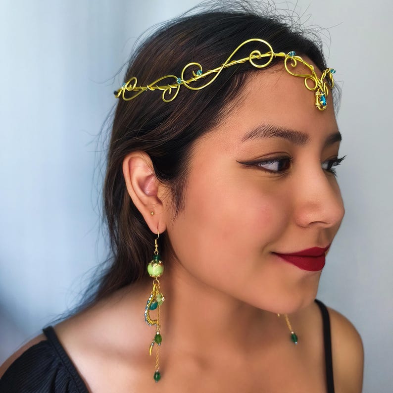 K&ouml;nnte beinhalten: Ein Kopfschmuck aus goldenem Draht mit zarten Schn&ouml;rkeln und kleinen blauen Kristallen, kombiniert mit einem langen, h&auml;ngenden Ohrring aus goldenem Draht, einer hellgr&uuml;nen marmorierten Perle und kleineren gr&uuml;nen Kristallen in gestufter Anordnung.
