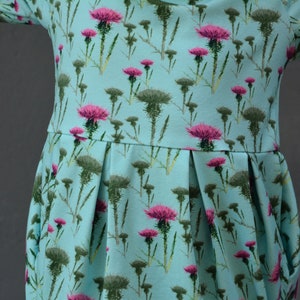 Puede incluir: Vestido azul claro con un estampado floral de cardos rosas y hojas verdes. El vestido tiene un corpi&ntilde;o ajustado, una falda plisada y mangas largas. La tela es un material suave y ligero, adecuado para ropa informal.