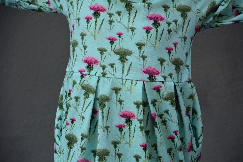 Puede incluir: Vestido azul claro con un estampado floral de cardos rosas y hojas verdes. El vestido tiene un corpi&ntilde;o ajustado, una falda plisada y mangas largas. La tela es un material suave y ligero, adecuado para ropa informal.