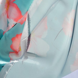 May include: A close-up of a light blue fabric with a white and pink floral print. The fabric is folded in half, showing the front and back sides of the print.