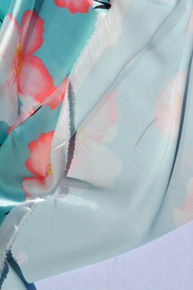 May include: A close-up of a light blue fabric with a white and pink floral print. The fabric is folded in half, showing the front and back sides of the print.