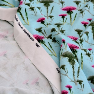 Puede incluir: Primer plano de tela con fondo azul claro y un patr&oacute;n repetido de flores de cardo rosas y follaje verde. La tela tiene un borde blanco con texto. La tela es probablemente de algod&oacute;n.