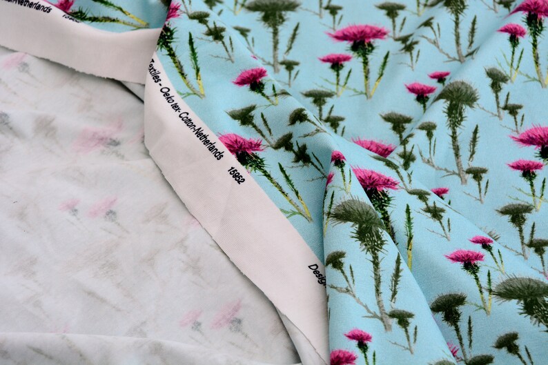 Puede incluir: Primer plano de tela con fondo azul claro y un patr&oacute;n repetido de flores de cardo rosas y follaje verde. La tela tiene un borde blanco con texto. La tela es probablemente de algod&oacute;n.
