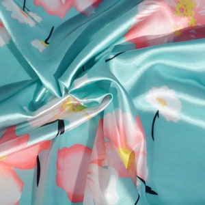 May include: A turquoise blue satin fabric with a floral print of pink and white flowers. The fabric is folded and draped, showing the texture and sheen of the satin.
