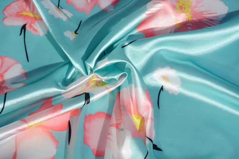 May include: A turquoise blue satin fabric with a floral print of pink and white flowers. The fabric is folded and draped, showing the texture and sheen of the satin.