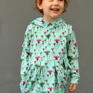 Puede incluir: Vestido con capucha azul claro con un estampado floral de cardos rosas y follaje verde. El vestido tiene mangas largas, una falda plisada y un corte holgado. El ni&ntilde;o sonr&iacute;e, usando el vestido sobre un fondo gris.