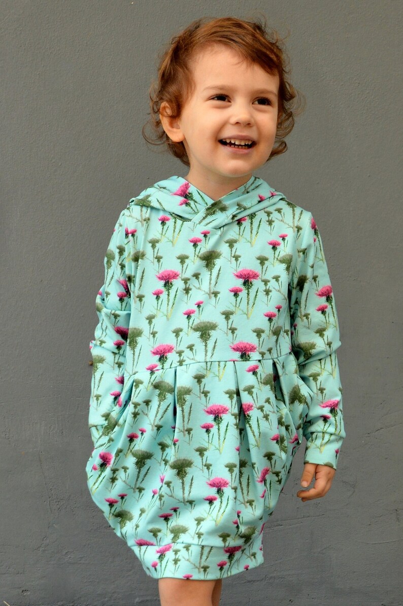 Puede incluir: Vestido con capucha azul claro con un estampado floral de cardos rosas y follaje verde. El vestido tiene mangas largas, una falda plisada y un corte holgado. El ni&ntilde;o sonr&iacute;e, usando el vestido sobre un fondo gris.