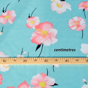 May include: A turquoise fabric with a pink floral print. The fabric is measured with a ruler in inches. The ruler reads "centimetres".