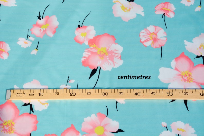 May include: A turquoise fabric with a pink floral print. The fabric is measured with a ruler in inches. The ruler reads "centimetres".