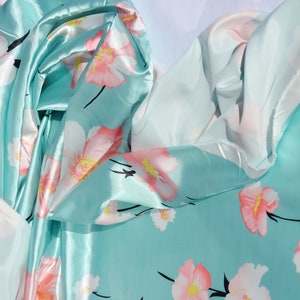 May include: A turquoise blue satin fabric with a floral print of pink and white flowers. The fabric is folded and draped, showing the texture and sheen of the satin.