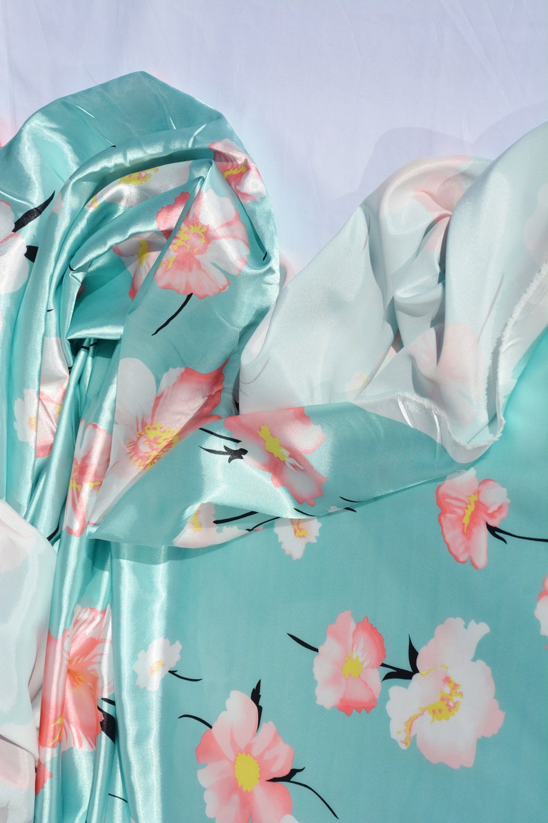 May include: A turquoise blue satin fabric with a floral print of pink and white flowers. The fabric is folded and draped, showing the texture and sheen of the satin.