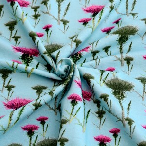 Puede incluir: Tela azul claro con un patr&oacute;n repetido de cardos. Los cardos tienen cabezas de color verde oscuro y flores de color rosa vibrante. La tela es probablemente de algod&oacute;n o un material similar, adecuado para ropa o decoraci&oacute;n del hogar.