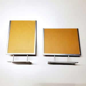 May include: Two metal sign holders with a silver frame and a tan backing. The sign holders have a rectangular shape and a base for standing. The background is white.