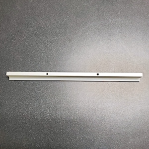 Op de afbeelding: Een lange, zilverkleurige metalen sierlijst met een decoratieve rand. De lijst heeft twee gelijkmatig verdeelde gaten en is ontworpen voor installatie. Het stuk wordt getoond tegen een donkergrijze achtergrond.