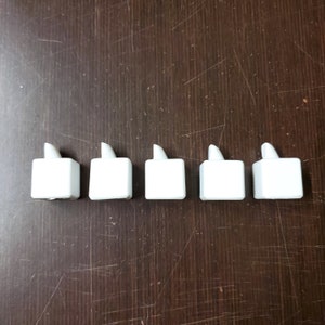 May include: Five white LED candle lights arranged in a row. Each candle has a square base and a flame-shaped top. The candles are on a dark brown wooden surface.