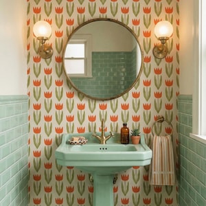 Op de afbeelding: Een badkamer in retrostijl met een mintgroene wastafel, gouden kranen en een ronde spiegel. Het behang heeft een herhalend patroon van oranje tulpen op een crèmekleurige achtergrond. Twee wandkandelaars met bolvormige lampen flankeren de spiegel.