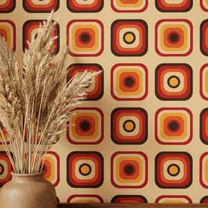 May include: A beige ceramic vase containing dried wheat stalks positioned against wallpaper featuring a repeating pattern of concentric rounded squares in shades of dark brown, red, orange, and yellow on a light tan background.