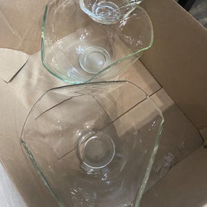 以下が含まれることがあります： さまざまなサイズの透明なガラス製サービングボウルのセット。ボウルは幾何学的な形状をしており、段ボール箱の中にあります。一番小さいボウルには金属製のハンドルが付いています。