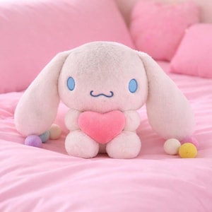 Puede incluir: Un conejo de peluche blanco con orejas largas, ojos azules y un corazón rosa. El peluche está sentado en una cama rosa con almohadas rosas. Pequeñas bolas de colores están esparcidas alrededor del conejo.
