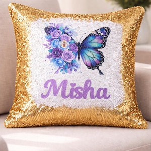May include: A square throw pillow covered in shimmering gold sequins. The center features a white rectangular panel displaying a blue and purple butterfly with a floral wing design and the name Misha printed in purple script lettering below it.