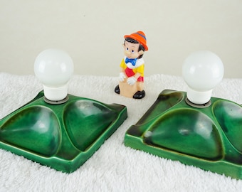 May include: Two green ceramic lamp bases with white light bulbs. The bases have a unique design with three rounded indentations. A small plastic figurine of a boy in a red and yellow shirt and a hat is standing in the center.