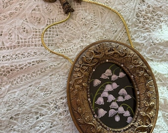 May include: An oval-shaped wall hanging featuring a dark center panel with a painted illustration of white lily of the valley flowers. The frame is textured gold with leaf motifs and small clear crystals, suspended by a gold cord with a brown bow.