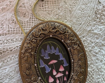 May include: An oval-shaped wall hanging featuring a dark center panel with painted blue bell-shaped flowers and pink mushrooms. The frame is metallic gold with intricate vine relief details, small embedded crystals, and a gold cord with a bow.