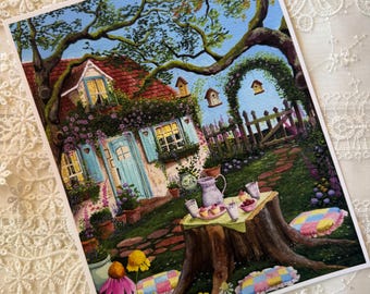 May include: A rectangular art print depicting a cottage with a red roof, blue shutters, and a stone path. A tree stump serves as a table with a pitcher, cups, and treats. The scene includes a garden with pink and yellow flowers, birdhouses, and colorful patchwork cushions on the grass.
