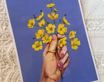 May include: A rectangular art print featuring a watercolor illustration of a hand holding a bouquet of bright yellow buttercup flowers against a solid periwinkle blue background. The hand has pink painted fingernails and a gold ring on the finger.