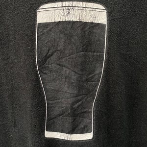 May include: Black sweatshirt featuring a graphic of a pint glass filled with a dark liquid. The pint glass design is outlined in white, with a white band at the top. The sweatshirt is made of a soft, textured fabric.