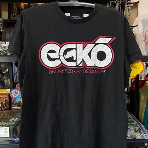 Może przedstawiać: Czarny T-shirt z białym logo "ecko" w czerwonym kółku. Tekst "UNLIMITED x BY DESIGN/®" znajduje się pod logo.