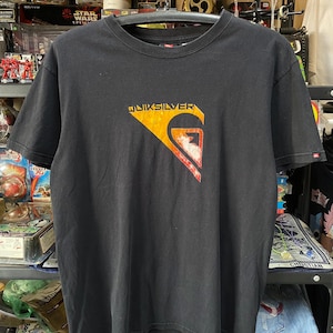 Pu&ograve; includere: T-shirt nero con un grande logo "QUIKSILVER" arancione e rosso sul davanti. Il logo &egrave; un'onda stilizzata con un fulmine al suo interno.