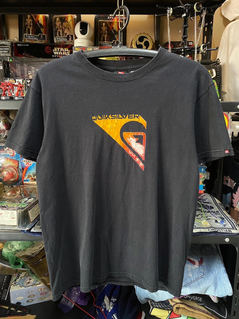 Pu&ograve; includere: T-shirt nero con un grande logo "QUIKSILVER" arancione e rosso sul davanti. Il logo &egrave; un'onda stilizzata con un fulmine al suo interno.