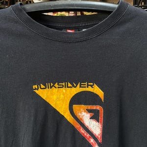 Pu&ograve; includere: Maglietta nera con un logo Quiksilver arancione e rosso sbiadito. Il logo &egrave; un'onda con una montagna sullo sfondo.