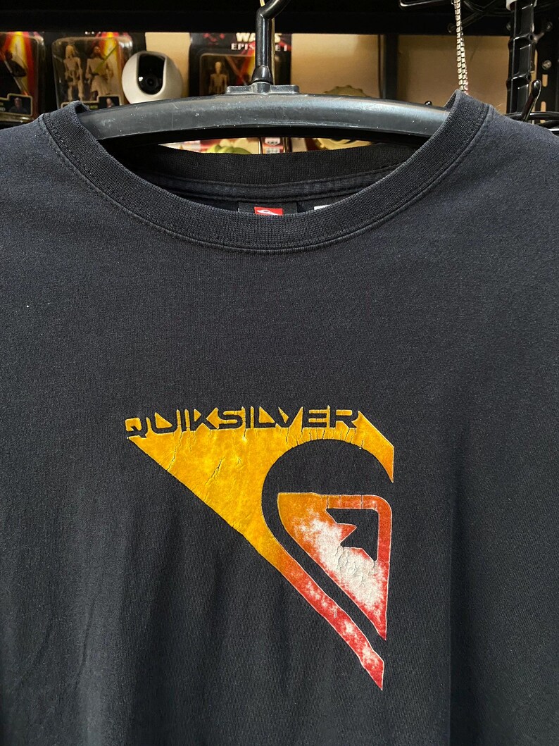 Pu&ograve; includere: Maglietta nera con un logo Quiksilver arancione e rosso sbiadito. Il logo &egrave; un'onda con una montagna sullo sfondo.