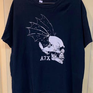 May include: Black t-shirt featuring a white graphic of a skull with bat wings. The text "A7X" is printed below the skull. The shirt has a crew neck and short sleeves.