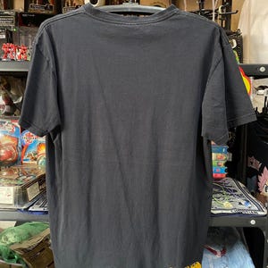 Pu&ograve; includere: Maglietta nera con un logo "Quiksilver" giallo in basso a destra. La maglietta &egrave; appesa a un appendiabiti.