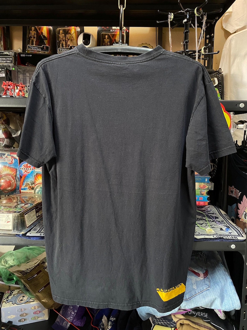 Pu&ograve; includere: Maglietta nera con un logo "Quiksilver" giallo in basso a destra. La maglietta &egrave; appesa a un appendiabiti.