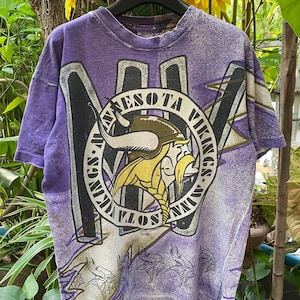 Puede incluir: Camiseta vintage de color morado, blanco y dorado con un gráfico de un casco vikingo y el texto "Minnesota Vikings".