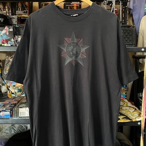 May include: A black t-shirt with a large starburst design on the front. The starburst has a central globe graphic with red and gray accents. The shirt is hanging on a wooden hanger.
