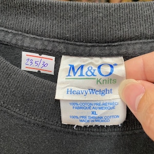 May include: Close-up of a black t-shirt collar with a tag. The tag reads "M&O Knits HeavyWeight" and includes text in English and French. The shirt also has a small white tag with the numbers "23.5/30".