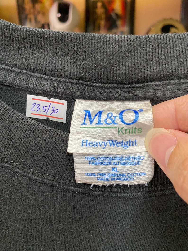 May include: Close-up of a black t-shirt collar with a tag. The tag reads "M&O Knits HeavyWeight" and includes text in English and French. The shirt also has a small white tag with the numbers "23.5/30".