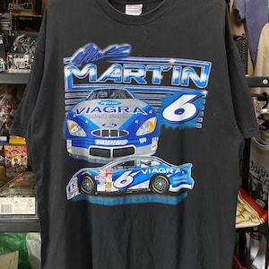 May include: Black t-shirt featuring a graphic of a race car with the text "Mark Martin 6" and the word "Viagra". The car is blue and silver with the Ford logo. The shirt is hanging on a black hanger.