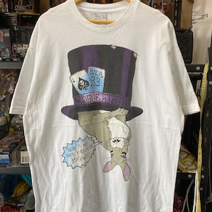 Può includere: T-shirt bianco con grafica di un coniglio con cappello a cilindro e carte da gioco. Il cappello ha la scritta "D.T.A. 33 ROGUE STREET". Un fumetto dice "THERE'S NO WHITE BIRD HERE BIG WIG".