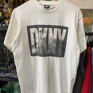May include: White t-shirt with a black and white graphic of the word "DKNY" with a cityscape in the background.